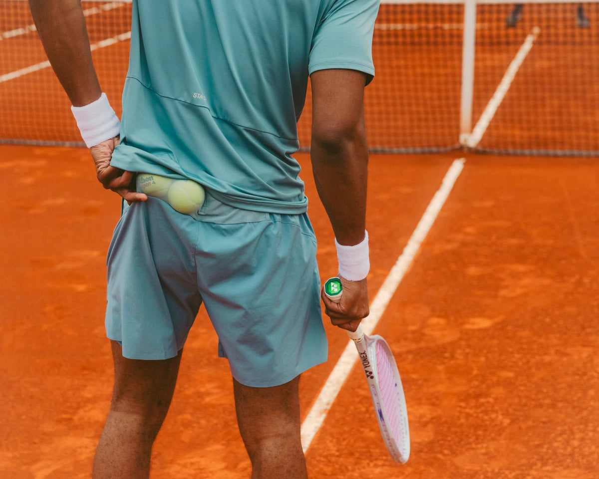 A tennis player on an orange clay court, shown from behind, wearing a light blue athletic shirt and shorts, holding a tennis racket and a tennis ball.